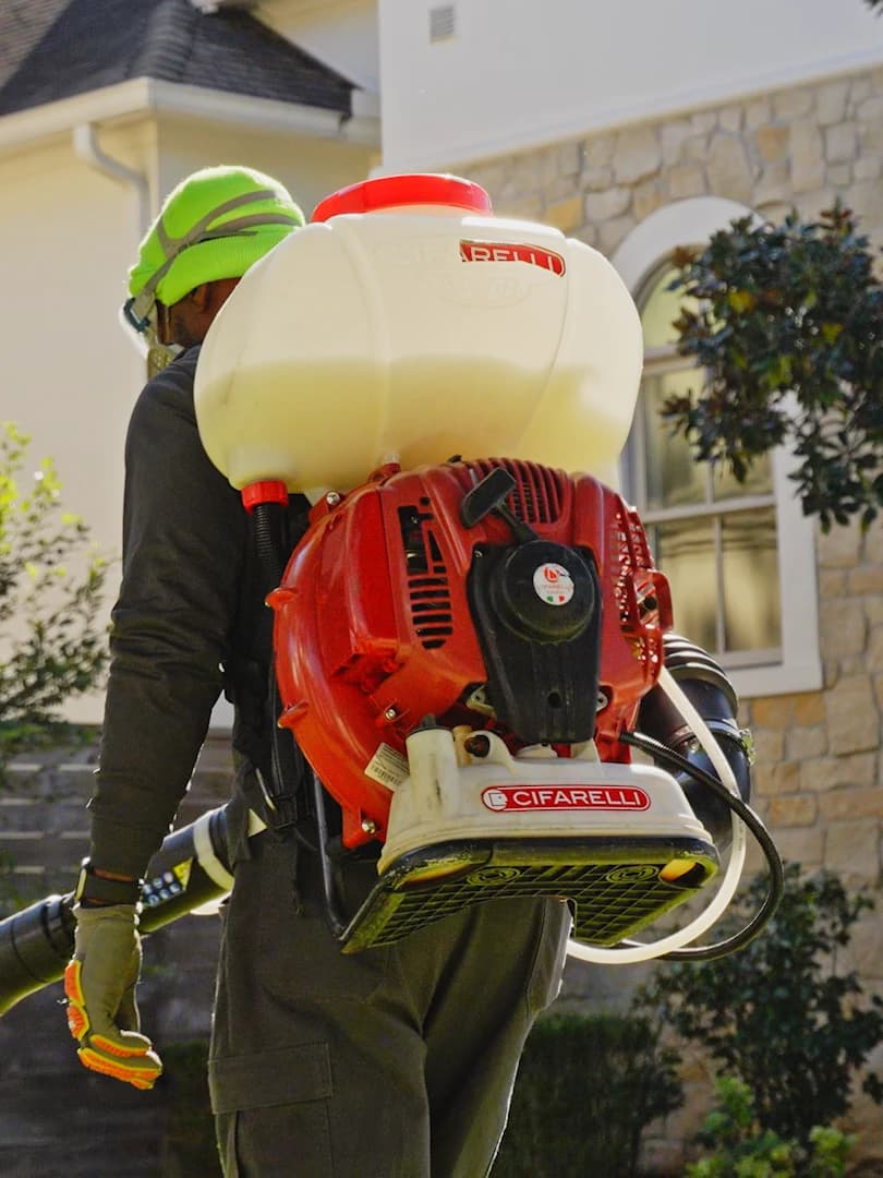 Technician wearing a backpack mist blower while treating the exterior of a stone-sided home